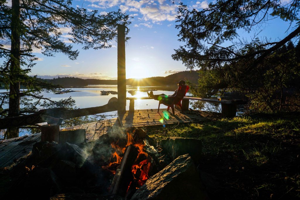 Woman looks over sea while the sun sets with a fire in the foreground