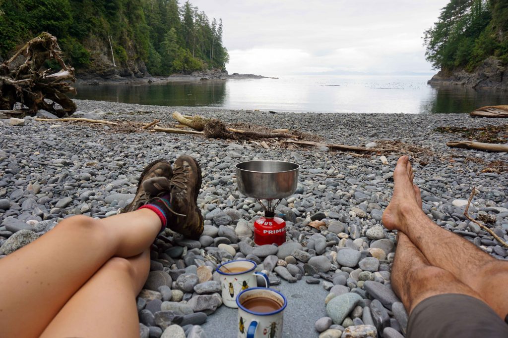 Two people making tea on a portable stove on a stone beach