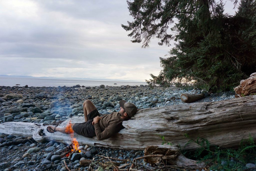 Man lies on beach log next to campfire