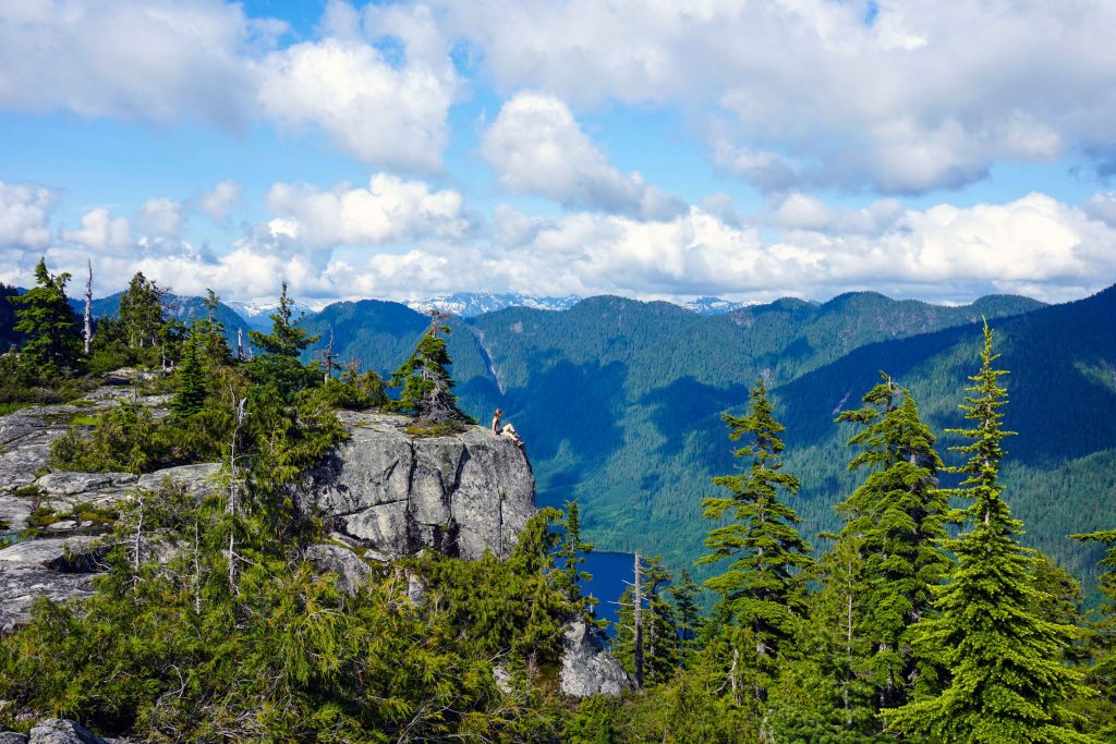 Woman sits on edge of cliff and looks over lake and tree covered mountains