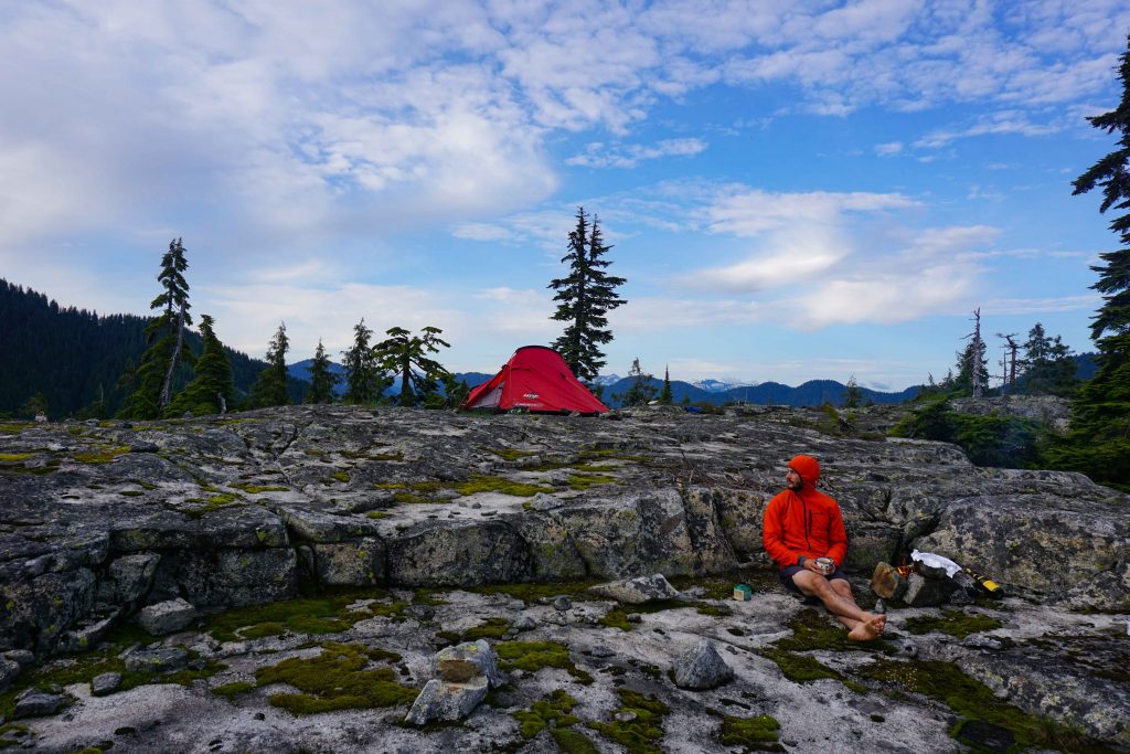 Man in orange top sits next to fire on rocky plateau with rent tent in the background