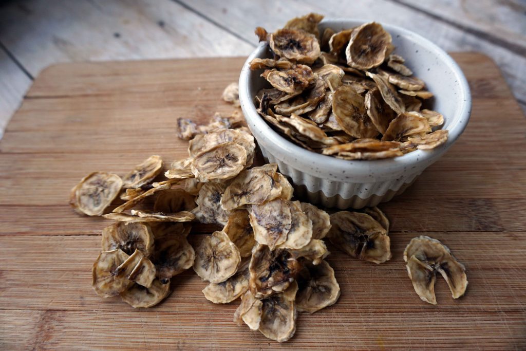 Dehydrated banana chips in a white bowl on a wooden chopping board