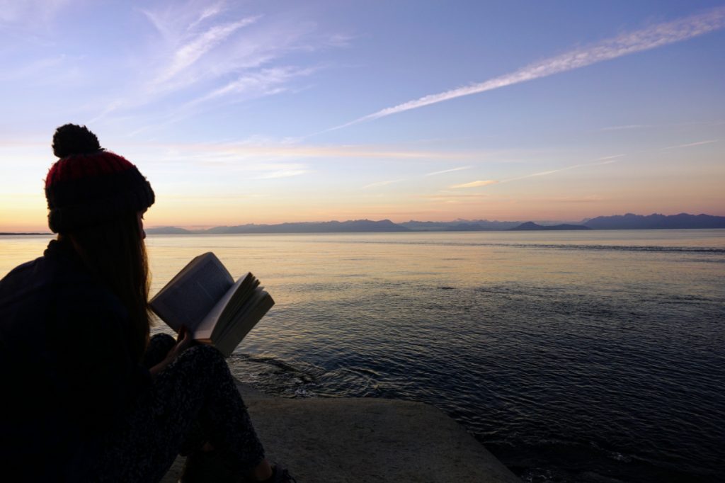 Woman reads book next to the ocean as the sun sets