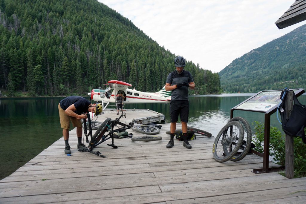 Two men dismantle bikes while standing on a wooden wharf on a lake with floatplane in the background