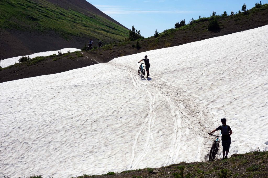 Mountain bikers walk bikes across a summer snow field on a mountain
