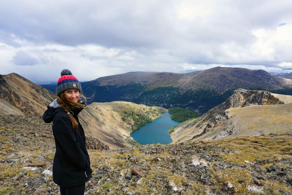 Woman on mountain ridge smiles at camera with blue alpine lake in the distance